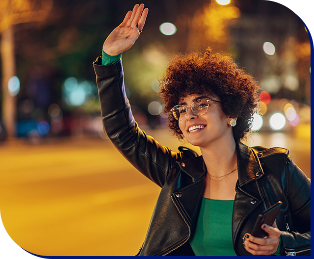 Passageira sorrindo com o braço levantado chamando uma corrida na rua à noite, com as luzes da cidade desfocadas atrás.